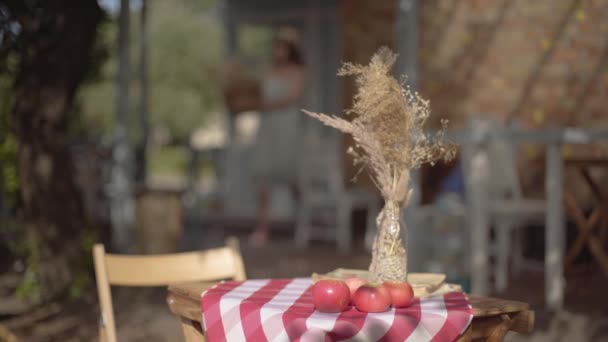Verschwommene Figur eines jungen Landmädchens, das mit dem Weidenkorb im Hintergrund geht. kleiner Tisch mit Äpfeln und Vase mit dekorativem Gras im Vordergrund. ländlicher Lebensstil — Stockvideo
