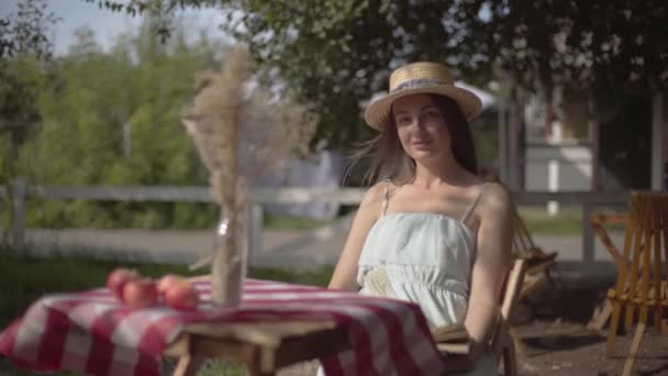 Bir hasır şapka ve beyaz elbiseli genç kırsal kız kitap okuma dekoratif çim ile vazo ile küçük bir masada oturuyor. Kırsal yaşam tarzı. Güzel bir yaz gününde boş zaman — Stok video