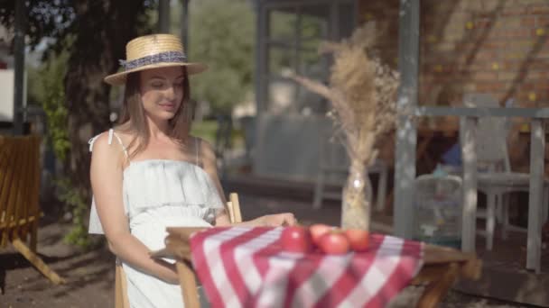 Młoda dziewczyna w słomkowym kapeluszu i białej sukni siedzącej przy małym stole i patrząc na kamerę. Wiejski styl życia. Wypoczynek w pięknym letnim dniu. — Wideo stockowe
