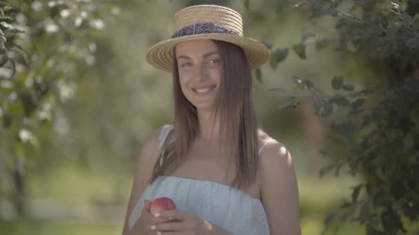 Ελκυστική νεαρή θετική γυναίκα με ψάθινο καπέλο και λευκό φόρεμα κοιτάζοντας την κάμερα χαμογελώντας κρατώντας μήλο στέκεται στο καταπράσινο καλοκαιρινό κήπο. Χρόνος συγκομιδής, αγροτική ζωή — Αρχείο Βίντεο