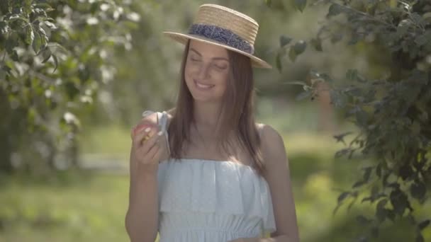 Adorable joven mujer positiva de moda en sombrero de paja y vestido blanco mirando a la cámara sonriendo sosteniendo manzana de pie en el verde jardín de verano. Tiempo de cosecha, estilo de vida rural — Vídeos de Stock