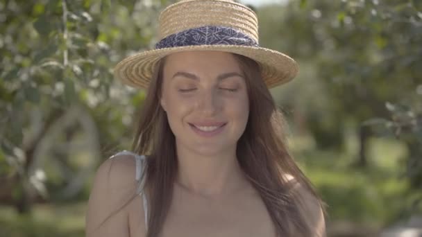 Чарівна молода жінка в солом'яному капелюсі і білій сукні дивиться на камеру, що стоїть в зеленому літньому саду. Сільський спосіб життя . — стокове відео