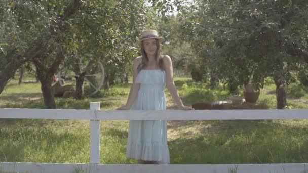 밀짚 모자와 긴 흰색 드레스를 입은 귀여운 젊은 여성이 울타리 뒤의 녹색 여름 정원에 서있는 카메라를 바라보고 있습니다. 농촌 생활 — 비디오