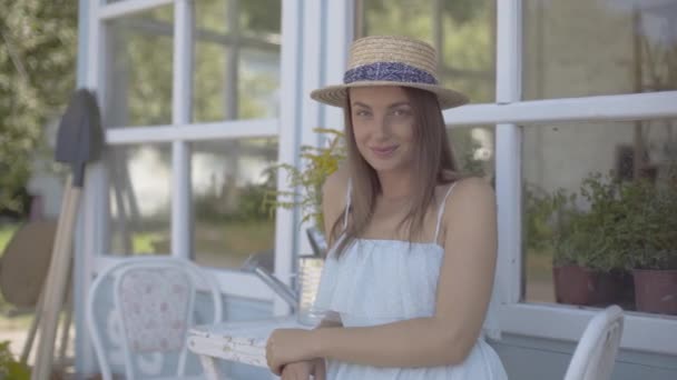 Mujer joven de moda en un sombrero de paja y vestido blanco sonriendo mientras huele flores silvestres en una regadera sentada en la silla frente a la pequeña casa del pueblo. Estilo de vida rural — Vídeo de stock