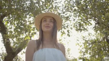 Yeşil yaz bahçesinde duran hasır şapka lı ve uzun beyaz elbiseli çekici genç kadın. Güneşin arkasında parlıyor. Kırsal yaşam tarzı. Yavaş çekim.