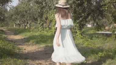 Hasır şapkalı ve yeşil yaz bahçesinde koşan uzun beyaz elbiseli çekici genç kadının geri görünümü sonra dönüp davetkar bir jest yapıyor. Tasasız kırsal yaşam. Yavaş çekim