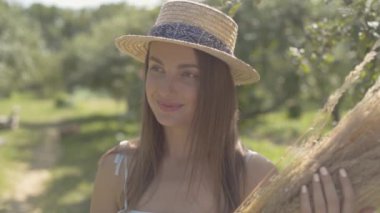 Hasır şapkalı ve uzun beyaz elbiseli çekici genç kadın, yeşil yaz bahçesinde gülümseyerek kameraya bakarak bir demet kuru ot tutuyor. Kırsal yaşam tarzı. Yavaş çekim