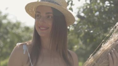 Hasır şapkalı ve uzun beyaz elbiseli sevimli genç kadın, yeşil yaz bahçesinde kuru ot demet tutan ayakta. Kırsal yaşam tarzı. Yavaş çekim.