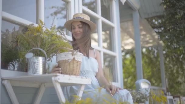 麦わら帽子と白いドレスを着た魅力的な若い女性は、水やりで野生の花を嗅ぎながら微笑みながら、小さな村の家の前の椅子に座ることができます。田舎のライフスタイル — ストック動画
