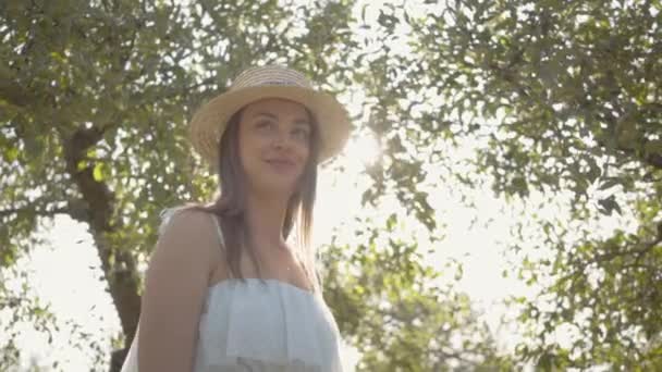 Όμορφη νεαρή γυναίκα με ψάθινο καπέλο και μακρύ λευκό φόρεμα στέκεται στο καταπράσινο καλοκαιρινό κήπο. Ο ήλιος λάμπει πίσω από το κορίτσι. Αγροτική ζωή. Αργή κίνηση. — Αρχείο Βίντεο