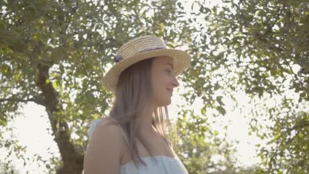 Carina giovane donna in cappello di paglia e lungo abito bianco in piedi nel verde giardino estivo. Il sole splende dietro la ragazza. Stile di vita rurale. Rallentatore . — Video Stock