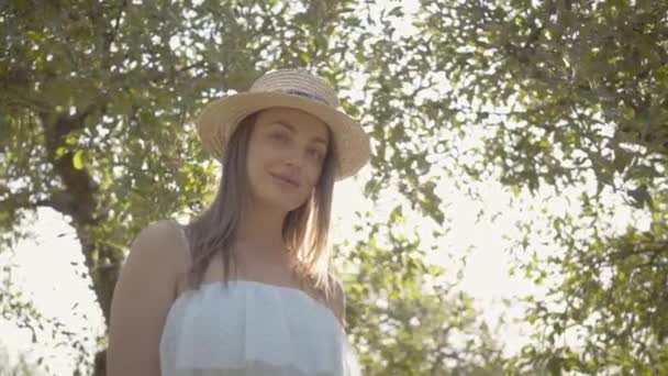 Привлекательная молодая женщина в соломенной шляпе и длинном белом платье смотрит на камеру, улыбаясь стоя в зеленом летнем саду. Солнце светит позади девушки. Сельский образ жизни — стоковое видео