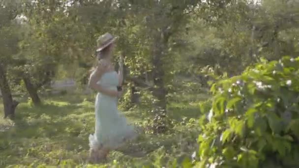 Imádnivaló, fiatal nő, szalma kalap és a hosszú, fehér ruha fut át a zöld nyári kert. Gondtalan falusi élet, kapcsolat a természettel. Lassított mozgás. — Stock videók
