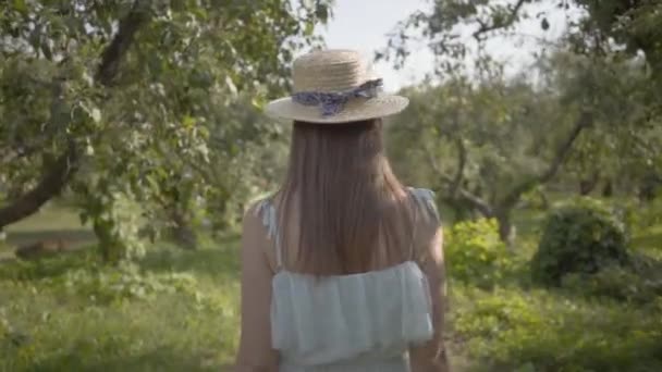 Vista posteriore di giovane donna carina in cappello di paglia e lungo abito bianco passeggiando attraverso il verde giardino estivo. Vita rurale spensierata, connessione con la natura. Rallentatore . — Video Stock