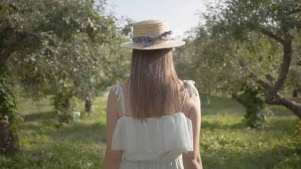 Vista posteriore di attraente giovane donna in cappello di paglia e lungo abito bianco passeggiando attraverso il verde giardino estivo poi girando e facendo gesto invitante. Vita rurale spensierata, connessione con la natura — Video Stock