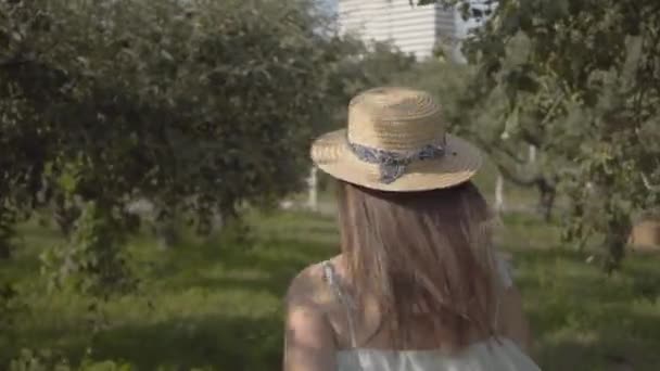 Vista posterior de la atractiva mujer joven en sombrero de paja y vestido blanco largo corriendo a través del verde jardín de verano. Vida rural despreocupada, conexión con la naturaleza. Movimiento lento . — Vídeo de stock