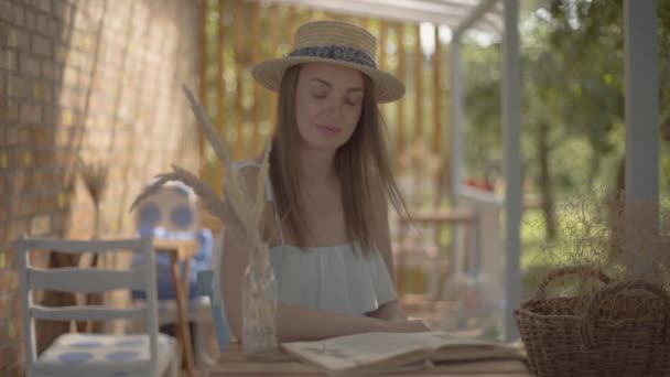 Bella ragazza rurale in un cappello di paglia e vestito bianco seduto al piccolo tavolo di legno all'aperto a leggere il libro. Stile di vita rurale. Tempo libero in una bella giornata estiva. Rallentatore . — Video Stock