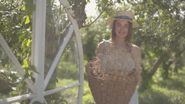 Милая молодая женщина в соломенной шляпе, держащая плетеную корзину с травами и глядя на камеру, улыбающуюся в зеленом летнем саду. Сельский образ жизни. Медленное движение — стоковое видео