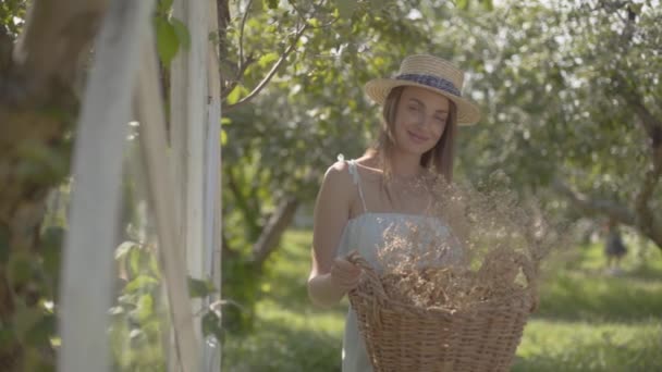 초록색 여름 정원에서 웃고있는 카메라를 보고 고리버들 바구니를 들고 있는 밀짚 모자를 쓴 예쁜 젊은 여성. 농촌 생활 방식. 슬로우 모션 — 비디오