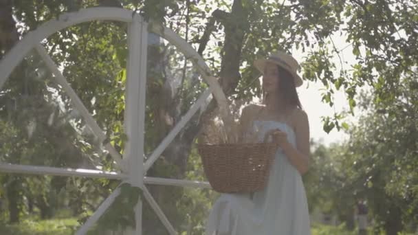 Schattige jonge vrouw in stro hoed het houden van de rieten mand met kruiden en kijken naar de camera glimlachend in de groene zomertuin. Landelijke levensstijl. Slow Motion — Stockvideo