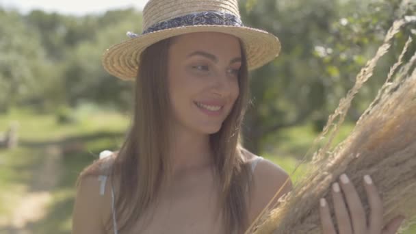 Mulher jovem bonito em chapéu de palha e vestido branco longo em pé no jardim de verão verde segurando um monte de grama seca. Estilo de vida rural. Movimento lento . — Vídeo de Stock