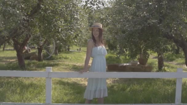 Jovem atraente em chapéu de palha e vestido branco longo olhando para longe e para a câmera sorrindo em pé no jardim de verão verde por trás da cerca. Estilo de vida rural. Movimento lento . — Vídeo de Stock