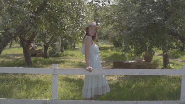 Модна молода жінка в солом'яному капелюсі і довга біла сукня, що стоїть в зеленому літньому саду за парканом. Сільський спосіб життя. Повільний рух . — стокове відео