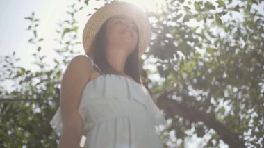 Hasır şapka lı ve beyaz elbiseli sevimli genç kadın elma ları toplayıp yeşil bahçedeki hasır sepetine koyuyor. Hasat zamanı, kırsal yaşam tarzı. Alt görünüm. Yavaş çekim