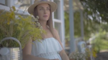 Küçük bir köy evinin önünde oturan bir hasır şapka ve beyaz elbise li büyüleyici genç kadın. Kırsal yaşam tarzı. Yavaş çekim.