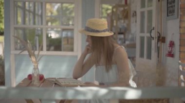 Genç kırsal kız bir hasır şapka ve beyaz elbise kitap okuma açık havada küçük ahşap masada oturan. Kırsal yaşam tarzı. Güzel bir yaz gününde boş zaman. Yavaş çekim.