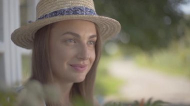 Küçük köy evinin önünde oturan hasır şapkalı ve beyaz elbiseli sevimli genç kadının yakın çekim yüzü. Kırsal yaşam tarzı. Yavaş çekim.