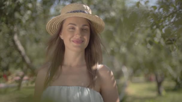 Retrato de moda jovem mulher em chapéu de palha e vestido branco longo olhando para a câmera sorrindo em pé no jardim de verão verde segurando um monte de grama seca. Estilo de vida rural. Movimento lento — Vídeo de Stock