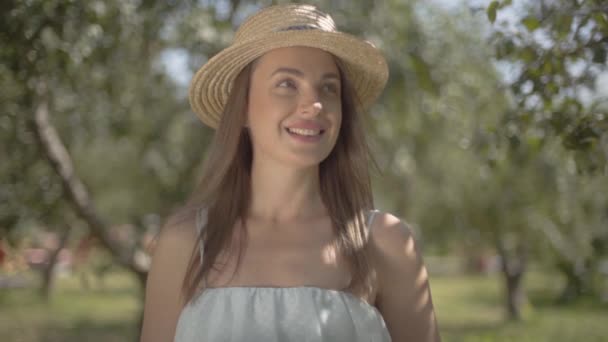 Eine junge Frau mit Strohhut und langem weißen Kleid blickt lächelnd in die Kamera, die im grünen Sommergarten steht und trockenes Gras in der Hand hält. ländlicher Lebensstil. Zeitlupe — Stockvideo