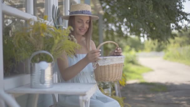 Attraente giovane donna in un cappello di paglia e vestito bianco mettendo piccolo cesto sul tavolo seduto di fronte alla piccola casa di villaggio. Stile di vita rurale. Rallentatore — Video Stock