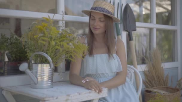 Mooie jonge vrouw in een stro hoed en witte jurk zittend in de voorkant van het kleine dorpshuis. Landelijke levensstijl. Slow Motion. — Stockvideo