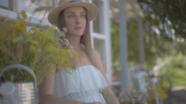 Affascinante giovane donna con cappello di paglia e vestito bianco seduta di fronte alla piccola casa di paese. Stile di vita rurale. Rallentatore . — Video Stock