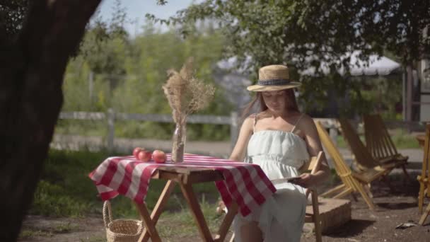 Menina rural jovem em um chapéu de palha e vestido branco sentado na pequena mesa com vaso com grama decorativa lendo o livro. Estilo de vida rural. Lazer em um belo dia de verão — Vídeo de Stock