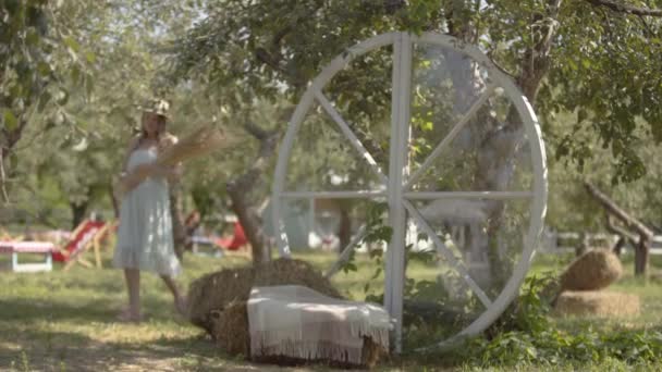 Attraktive junge Frau mit Strohhut und langem weißen Kleid im grünen Sommergarten mit einem Strauß Gras. ländlicher Lebensstil. — Stockvideo