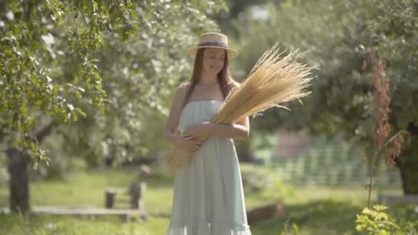 Мила молода жінка в солом'яному капелюсі і довга біла сукня дивиться на камеру, посміхаючись стояти в зеленому літньому саду, тримаючи купу трави. Сільський спосіб життя . — стокове відео