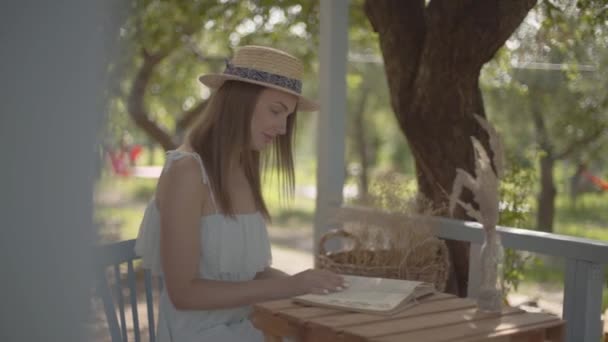 Mooie jonge landelijke meisje in een stro hoed en witte jurk zittend op de kleine houten tafel buiten het lezen van het boek. Landelijke levensstijl. Vrije tijd op een mooie zomerdag. Slow Motion. — Stockvideo