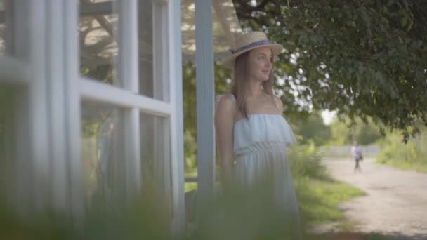 小さな村の家の前で野生の花を嗅ぎながら、麦わら帽子と白いドレスを着たかわいい若い女性。田舎のライフスタイル。スローモーション. — ストック動画
