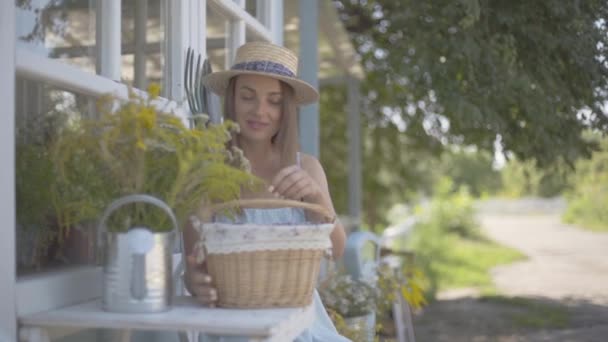 Schattige jonge vrouw in een stro hoed en witte jurk het nemen van Apple uit een kleine mand zittend in de voorkant van het kleine dorpshuis. Landelijke levensstijl. Slow Motion — Stockvideo