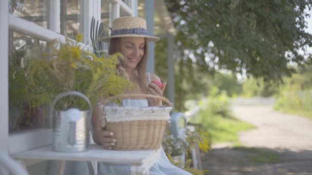 Hermosa joven con un sombrero de paja y vestido blanco tomando manzana de una pequeña canasta sentada frente a la pequeña casa del pueblo. Estilo de vida rural. Movimiento lento — Vídeos de Stock