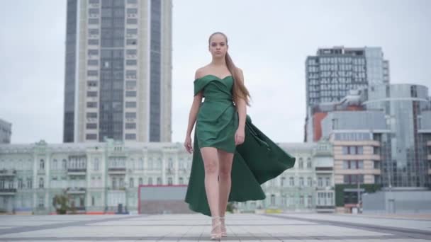 Hermosa chica hermosa en un vestido verde de noche impresionante caminando en la plaza vacía de la ciudad cerca de rascacielos. Serie de personas reales. Movimiento lento — Vídeo de stock