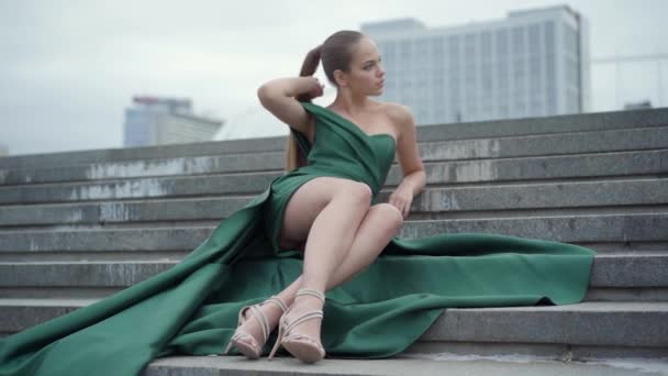 Schattig prachtig meisje in een prachtige avond groene jurk zittend op trappen fascinerend op lege stadsplein in de buurt van wolkenkrabber. Echte mensen serie. Slow Motion — Stockvideo