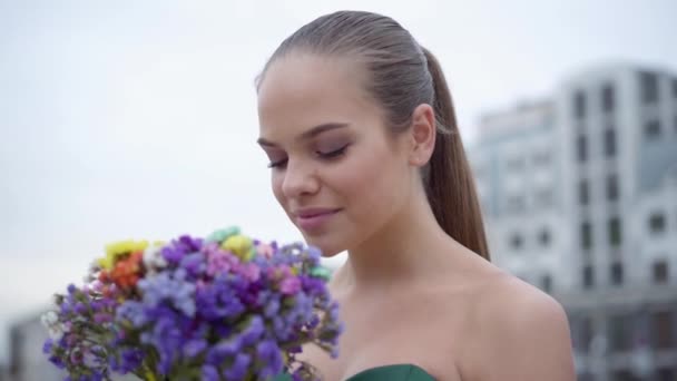 Portret van een schattig meisje in chique jurk ruikende een geurige boeket van wilde bloemen op de achtergrond van de ochtend stad. Echte mensen serie. — Stockvideo