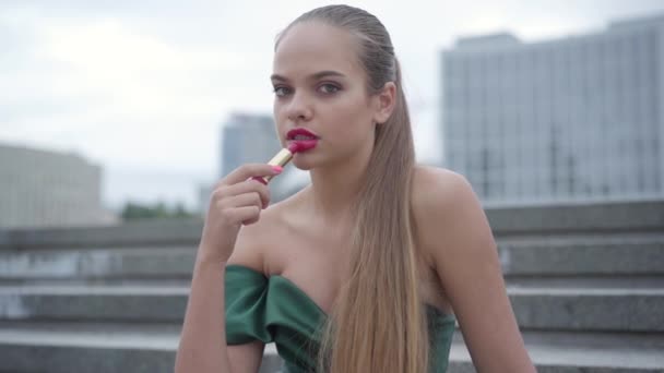 Portrait of beautiful young girl in emerald dress painting lips with bright pink lipstick on the background of a morning city. Real people series. Slow motion — Stock Video