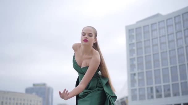 Piękna Wspaniała dziewczyna w oszałamiającej wieczornej, zielonej sukni tańczy fascynująco na pustym placu miejskim w pobliżu wieżowca. Seria prawdziwych ludzi. Zwolnionym — Wideo stockowe