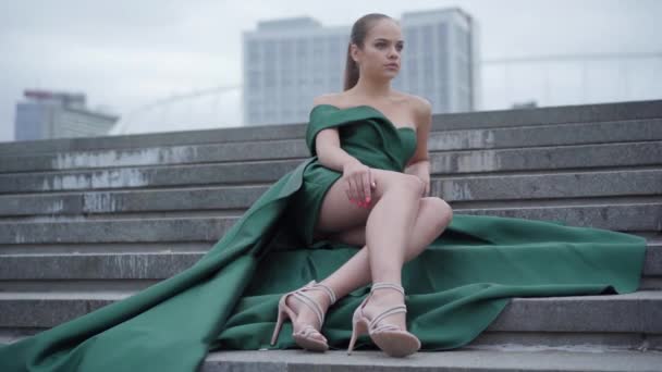 Hermosa chica hermosa en un vestido de noche verde impresionante sentado en las escaleras fascinantemente en la plaza vacía de la ciudad cerca de rascacielos. Serie de personas reales . — Vídeo de stock