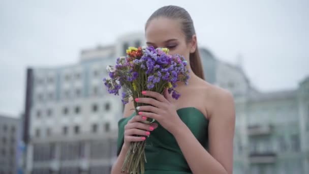 Красивая девушка в шикарном платье, стоящая с ароматным букетом полевых цветов на фоне утреннего города. Реальные люди . — стоковое видео